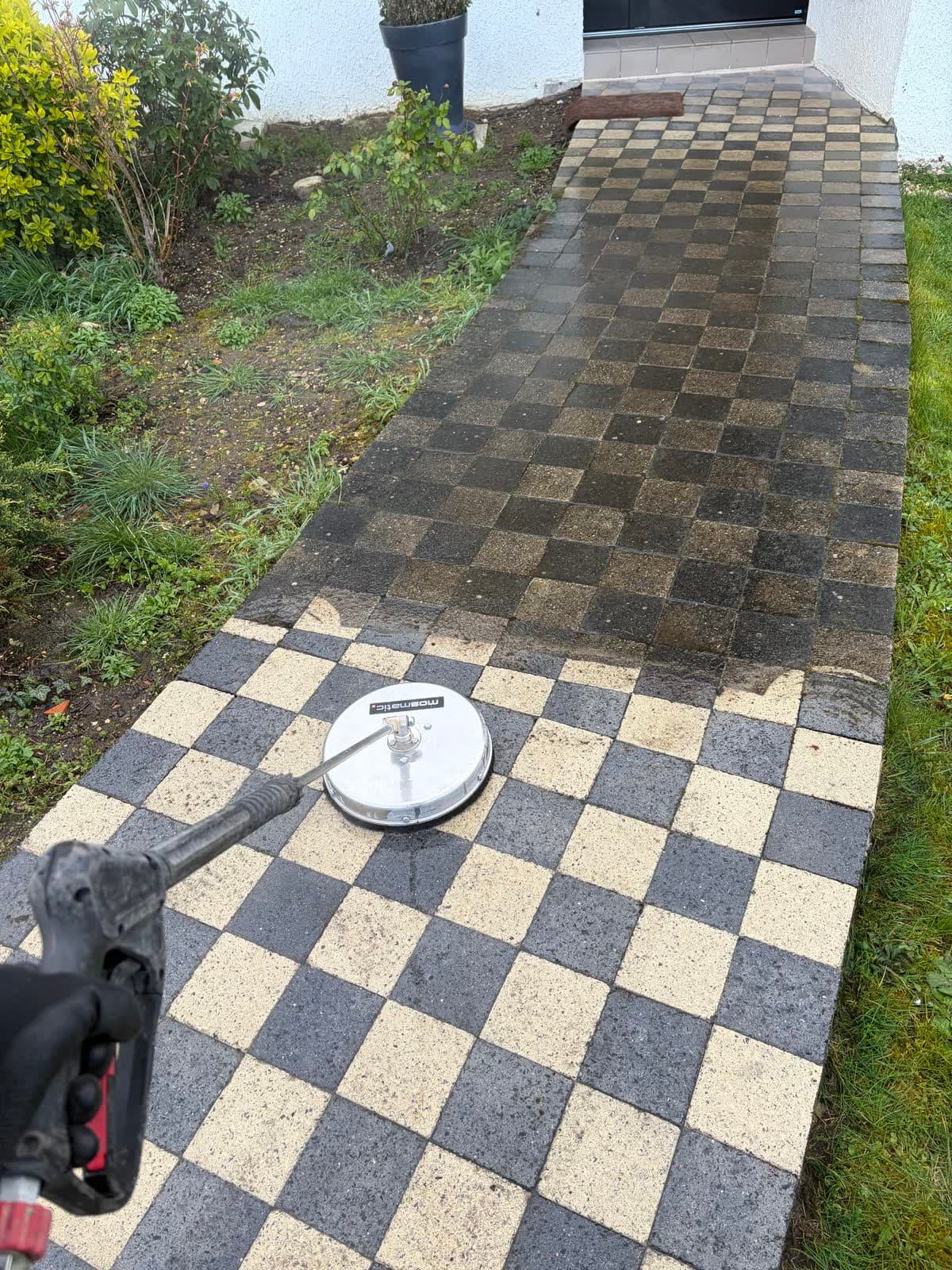 Allée en pavés damier en cours de nettoyage : pavés encore noircis au fond, motif noir et beige retrouvé au premier plan